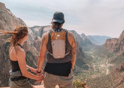 Jake Kramer and his wife hold hands on a canyon overlook in Zion National Park, the valley winding below.