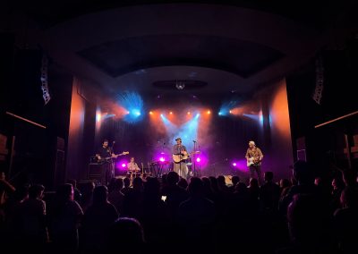 Jake Kramer plays a black bass guitar on stage right as blue and orange beams sweep over the crowd.