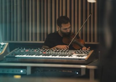 Jake Kramer records violin in a studio, bow raised above a synthesizer at the mixing desk.