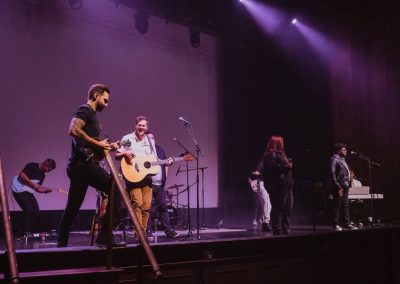 Jake Kramer plays mandolin at the front of a theater stage alongside an acoustic guitarist and vocalists.